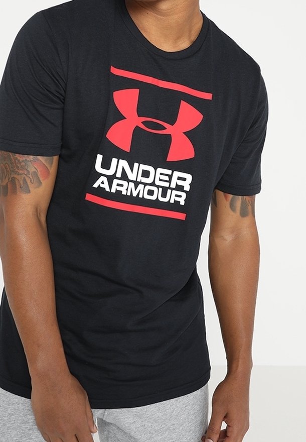 Hombre vistiendo camiseta negra de Under Armour con logo rojo y texto blanco, combinada con pantalones gris claro, de pie frente a un fondo blanco.