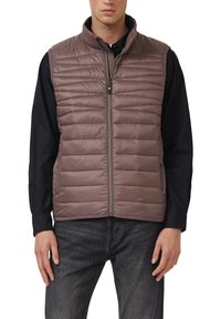 Veste matelassée taupe avec col montant, fermeture éclair et motif matelassé horizontal. Portée avec un chemisier noir à manches longues et un jean foncé.