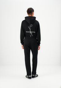 Black hoodie with a drawstring, featuring white text design on the back; paired with black pants and dark sandals.