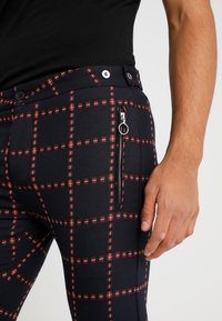 Marineblauwe geruite broek met een rood-witte ruitpatroon; heeft een zijritszak en twee knopen in de taille voor het vastmaken.