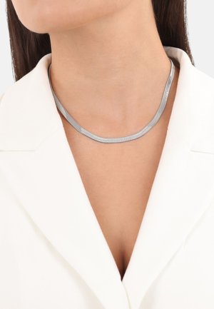 Collana choker in metallo argentato lucido, caratterizzata da un design elegante e flessibile con un sottile motivo a spina di pesce. Indossata su un indumento bianco.