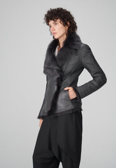 Chaqueta de cuero negra con cuello y ribete de pelo sintético, cremallera asimétrica y silueta estructurada, combinada con pantalones negros holgados.
