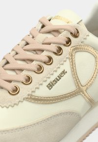 Baskets en cuir crème avec accents en daim, lacets beige et œillets dorés. Présente un logo cousu et des éléments de design courbes.