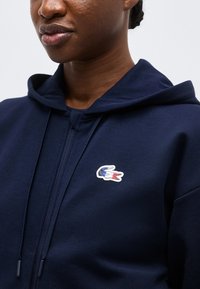 Inimese lähivaade, kes kannab tume sinist lukuga kapuutsi ja tri-värvilise krokodillilogoga vasakul rinnal.