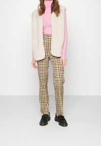 Cremefarbene Kunstpelzweste über einem rosa Rollkragenpullover, kombiniert mit grün-braunen karierten Hosen und schwarzen Schuhen mit flacher Sohle.