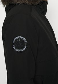 Black winter jacket with a round "Superdry Everest Expedition Company" patch on the upper sleeve and fur-lined hood.