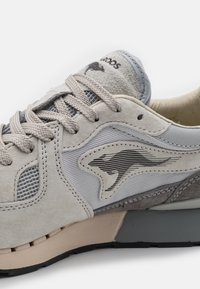 Grå joggesko med semsket skinn og mesh-detaljer, med en svart og grå logo, teksturerte lisser og en lett gummisåle.