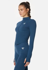 Chaqueta deportiva azul con cremallera fabricada en un tejido suave y elástico. Presenta mangas largas y un cuello alto, combinada con mallas a juego.