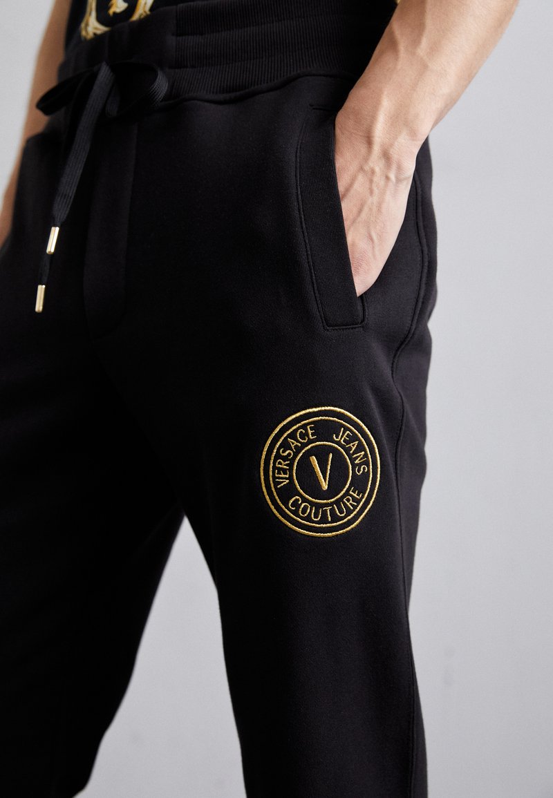 Pantalones de chándal negros con una cintura acanalada, que presentan un detalle de logotipo bordado en oro en el muslo izquierdo y bolsillos laterales.