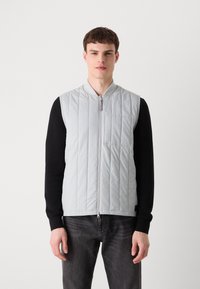 Colete acolchoado cinza claro, com fecho éclair e gola canelada. Design sem mangas, combinado com uma camisa preta de manga longa e jeans escuros.