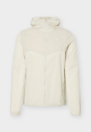 Světlá béžová bunda s kapucí, plným předním zipem, dlouhými rukávy a malým bílým logem Nike na levé straně hrudi.