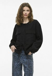 Čierna, dlhá bunda s dlhým rukávom a zapínaním na gombíky, s dvoma prednými vreckami. Vyrobená z texturovanej látky s nastaviteľným pásom. V spojení s modrými nohavicami s leopardím vzorom.