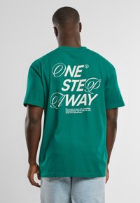 Camiseta de algod�ón verde con un corte holgado. La parte trasera cuenta con un texto blanco en negrita: "UN PASO MÁS CERCA" junto con frases motivacionales.