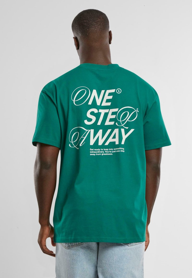 Camiseta de algodón verde con un corte holgado. La parte trasera cuenta con un texto blanco en negrita: "UN PASO MÁS CERCA" junto con frases motivacionales.