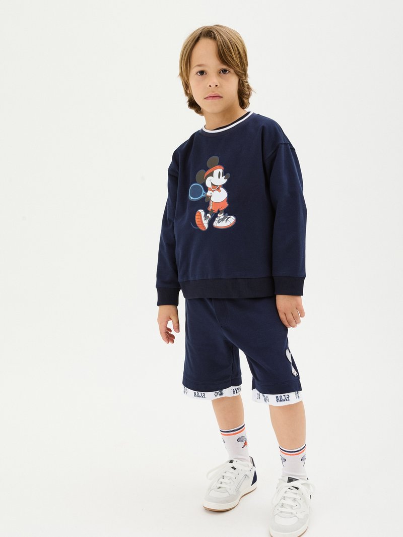 Dunkelblaues Sweatshirt mit weißem Kragen, das einen Cartoon-Charakter zeigt, kombiniert mit dunkelblauen Shorts und weißen Sneakers mit blauen Akzenten sowie gemusterten Socken.