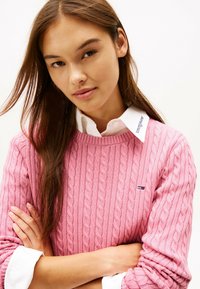 Maglione rosa a coste con colletto a coste, indossato sopra una camicia bianca con logo sul colletto. Braccia incrociate davanti.