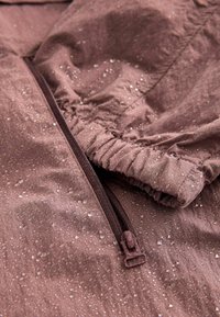 Tessuto di giacca marrone resistente all'acqua con dettagli in zip, polsini arricciati e gocce d'acqua sulla superficie, che evidenziano texture e design.