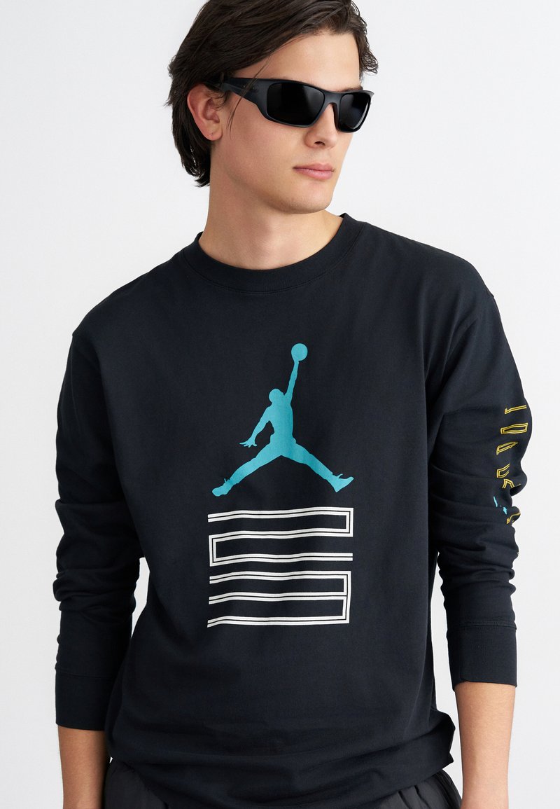 Mustas pikkade varrukatega särk, millel on tebalise siluett hüppavast figuurist ja valged horisontaalsed jooned. Varrukal on kollane teksti detail.