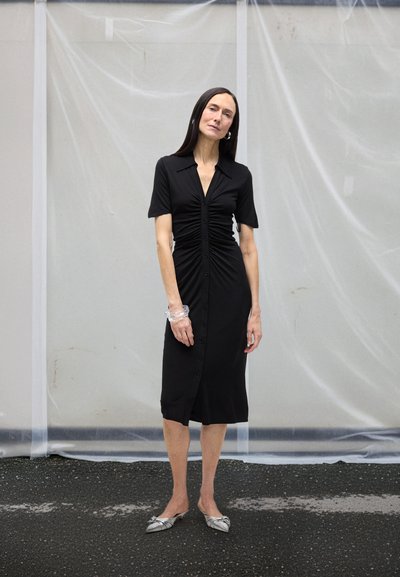 Woman with long dark hair wears a ruched black button-down dress, silver pointed flats, and a clear bracelet, standing on asphalt by a translucent wall.