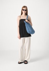 Blå denim slouchy veske, svart heklet tank topp, løse hvite bukser og svarte plattformsandaler. Modellen har på seg mørke solbriller og står.
