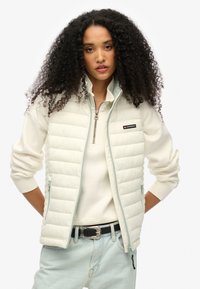 Weiß gepolsterte Jacke mit einem Reißverschluss an der Vorderseite, hohem Kragen und hellblauem Besatz. Hat ein Logopatch und gerippte Bündchen. Trägt man über einem hellen Oberteil.