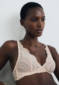 Bralette rendado em tom nude com borda recortada, apresentando padrões geométricos intrincados e alças ajustáveis. Textura suave, design leve.