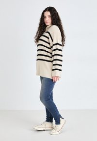 Beige and black striped oversized sweater paired with blue skinny jeans and white sneakers, featuring a relaxed fit and side slits.