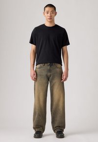 Μαύρο κοντομάνικο μπλουζάκι με άνετο, ξεθωριασμένο μπεζ τζιν παντελόνι. Το μοντέλο φοράει μαύρα παπούτσια. Απλό φόντο.