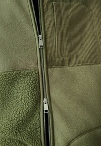 Olivengrøn jakke med en kombination af fleece- og ribfløjlsteksturer, lynlås i lukningen og en lodret ribfløjlslomme på brystet.