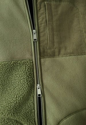 Olivengrøn jakke med en kombination af fleece- og ribfløjlsteksturer, lynlås i lukningen og en lodret ribfløjlslomme på brystet.