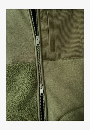 Olivovo zelená bunda kombinujúca textúry flísu a manšestru, so zipsovým zapínaním a vertikálnym manšestrovým vreckom na hrudi.