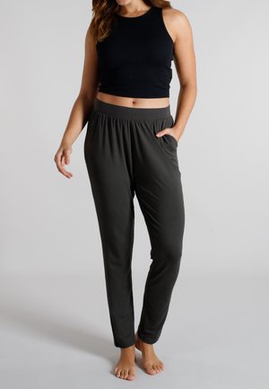 Frau trägt ein schwarzes ärmelloses Crop-Top und dunkle, graue Jogginghosen mit elastischem Taillenbund, steht barfuß und hat eine Hand in der Tasche.