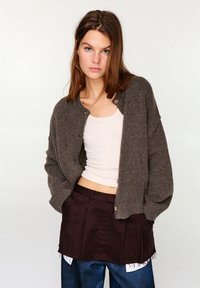 Bruine gebreide cardigan met lange mouwen, geribbelde manchetten en een ronde hals, met zichtbare knopen. Draagt over een beige tanktop en een donkere rok.
