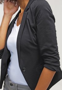 Musta bleiseri, jossa on kapea leikkaus, yksirivinen nappilista ja käärityt hihat valkoisen paidan päällä. Materiaali vaikuttaa kevyeltä ja rakenteiselta.