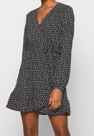 Vrouw in een zwarte wikkeljurk met lange mouwen, kleine witte bloemenprint en ruches aan de zoom, staand tegen een effen achtergrond.