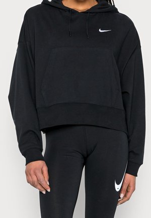 Persoon draagt een zwarte Nike-hoodie en zwarte legging met witte Nike swoosh-logo's, staand tegen een effen achtergrond.