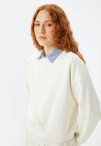 Lichtcrème sweatshirt met een ronde zoom, met daaronder een blauw-wit gestreept overhemd met kraag. Zachte textuur, relaxte pasvorm.