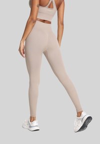 Žena u bež bezšavnim tajicama i odgovarajućim sportskim brusom, stoji s jednom nogom blago savijenom, u bijelim tenisicama na svijetlo sivoj pozadini.