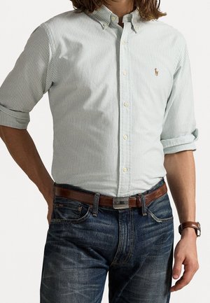 Mann trägt ein hellgrün gestreiftes Hemd mit Knöpfen und hochgekrempelten Ärmeln, blaue Jeans, braunen Ledergürtel und eine braune Armbanduhr und steht mit der Hand in der Hüfte.