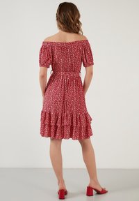 Femme debout de dos, portant une robe rouge à fleurs épaules dénudées avec ourlet à volant et des sandales rouges à talons bloc.