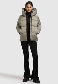 Pufjakke i afdæmpet grøn, med en stor hætte, lynlås foran og to sidelommer. Båret over en sort turtleneck og flagrende bukser.