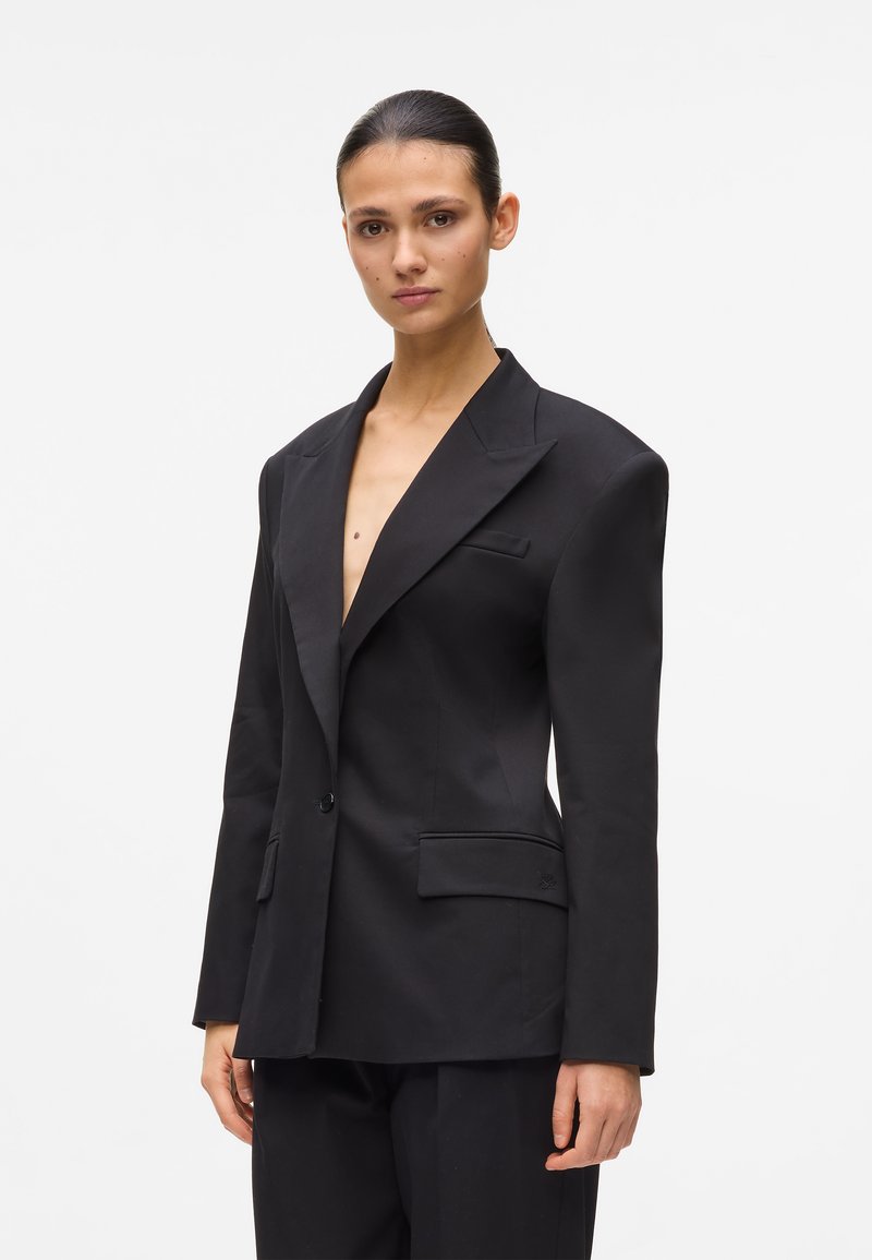 Γυναίκα με μαλλιά που έχουν χτενιστεί προς τα πίσω, φοράει ένα υπερμεγέθες μαύρο σακάκι με κοφτερές πέτες και ταιριαστό μαύρο παντελόνι, πάνω σε λευκό μονόχρωμο φόντο.