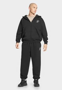 Czarna bluza z kapturem na zamek błyskawiczny z troczkami i kieszeniami z przodu, zestawiona z dopasowanymi czarnymi spodniami dresowymi z logo na udzie.