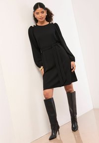 Černé šaty s dlouhými rukávy, stahováním v pase a ozdobnými detaily na ramenech, doplněné černými kozačkami na vysokém podpatku sahajícími ke kolenům, na modelce před neutrálním pozadím.