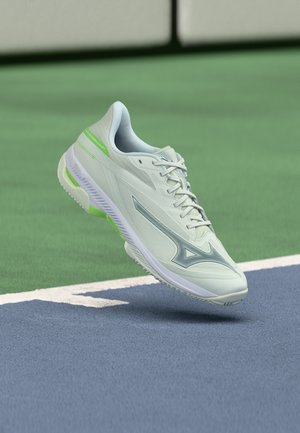 Világoszöld és fehér sportcipő logóval, a kék-zöld teniszpálya felszíne fölött lebeg, a határvonalak közelében.