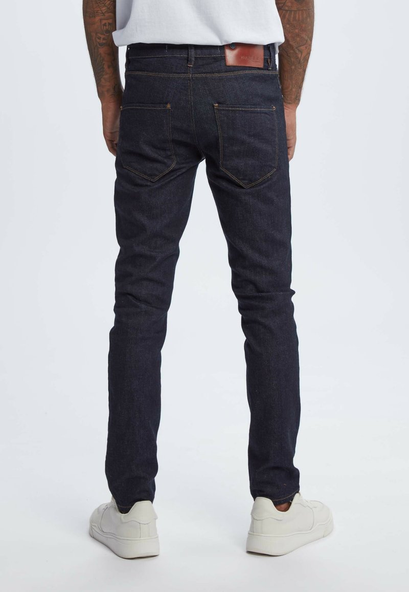 Personne portant un jean slim bleu foncé, des baskets blanches et une chemise blanche, se tenant de dos sur un fond clair uni.