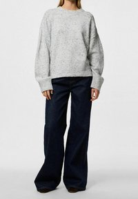 Personne portant un pull gris clair moucheté à manches longues et un pantalon large foncé, debout devant un fond uni.