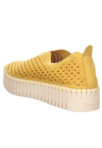 Scarpa gialla slip-on con tomaia perforata, dettaglio a onde e suola piatta beige texturizzata. Punta arrotondata e colletto imbottito.