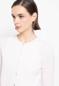 Cardigan bianco in tessuto morbido, con scollo rotondo e piccoli bottoni sul davanti. La texture appare liscia e leggera.