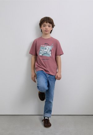 Camiseta gráfica rosa con diseño de calavera de dibujos animados y huesos cruzados. Combinada con jeans azules y zapatillas marrones. Fondo minimalista.